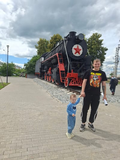 A young boy and a young man are posing for a photo in front of a decommissioned steam locomotive in Yanaul, Russia. The boy, wearing a blue long-sleeved shirt and blue pants, stands to the left of the man and holds his hand. The man, wearing a black t-shirt with a cartoon graphic and black pants with white stripes, is on the right, holding a rolled-up white object in his right hand. Both are looking at the camera and smiling. The steam locomotive is black with red accents, including the wheels and some piping, and features a prominent red star on its front. The ground is paved with grey interlocking bricks, and gravel is visible next to the train tracks. In the background, there are trees, buildings, and a cloudy sky. Another person in a grey uniform and a backpack is walking away from the camera on the right side of the image. The scene appears to be in a park or an outdoor museum dedicated to railway history.