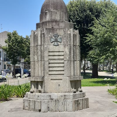 The image shows a close-up of a stone monument in Vila Verde, Portugal. The monument is a tall, tiered structure made of rough-hewn stone blocks. It has a domed top and is decorated with a cross emblem and horizontal ribbed patterns. In the foreground, there are green plants and white flowers. In the background, there is a street with cars, buildings, and trees, indicating an urban setting. The sky is clear and blue.