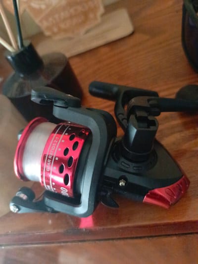 A fishing reel with a red and black body and white line is placed on a wooden surface. The reel has the inscription "GEAR RATIO 5.2:1". In the background, a blurred object that appears to be a bottle with reeds is visible. The scene is indoors, likely in a domestic setting, and there is no indication of the specific location being Zalesovo, Russia.