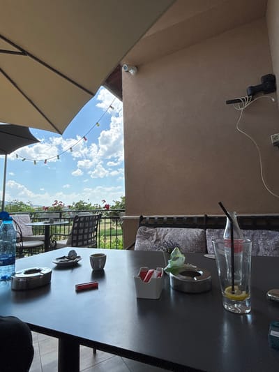 The image depicts an outdoor seating area at a cafe or restaurant, likely in the city of Ognyanovo, Bulgaria, given the context. The foreground features a dark, polished table with several items on it. These include an ashtray, a small dish with what appears to be a cigarette butt, a cup, a red rectangular object (possibly a lighter or matchbox), a white container with red and white packets (likely sugar and artificial sweetener), another ashtray, and a tall glass with a straw and a slice of lemon. To the left, a partially visible blue water bottle with a blue cap is present.
In the mid-ground, chairs and small tables are visible, set against a backdrop of lush greenery and flowers. A metal railing separates the seating area from the landscape beyond. The sky is a bright blue with scattered white clouds.
Overhead, a large umbrella provides shade, and decorative string lights with colored bulbs are strung across the scene. The architecture in the background suggests a building with a textured, light brown facade, and a security camera is mounted on the wall. The overall impression is of a relaxed, outdoor dining experience on a sunny day.