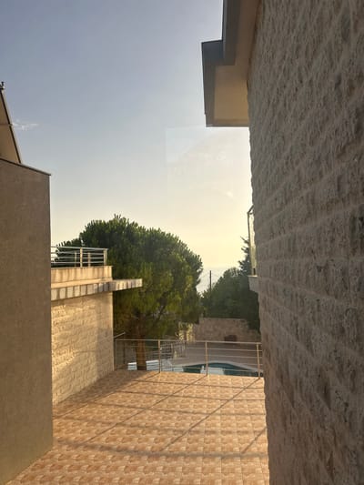 The image depicts a view of a tiled patio and a swimming pool area, framed by the exterior walls of buildings. On the left, a beige-colored building wall with a rough texture occupies the foreground. To the right, a wall constructed from light-colored stone blocks is prominently featured. In the middle ground, a tiled patio with a diamond pattern is visible, leading towards a swimming pool with blue water. A metal railing surrounds the pool area. A large green tree with dense foliage is situated behind the patio. In the background, more trees and distant buildings are present, with the sea and a hazy sky visible on the horizon. The setting appears to be a residential property in Brca, Montenegro, bathed in the warm light of either sunrise or sunset, casting long shadows across the patio. The overall scene conveys a sense of tranquility and a resort-like atmosphere.