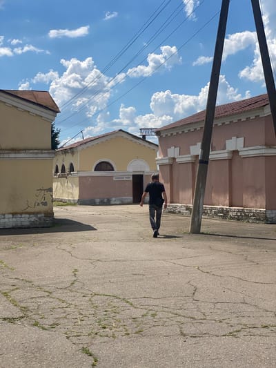 The image shows a man walking away from the camera on a cracked asphalt ground in the city of Romodan, Ukraine. He is wearing a black t-shirt and dark pants, with a dark bag slung over his shoulder. In the background, there are two low-rise buildings. The building on the left is painted yellow with a red roof and has arched windows. The building in the center, also yellow with a red roof, has a sign that reads "ЧОЛОВІЧИЙ ТУАЛЕТ" (Men's Toilet). To the right is a pink building with a red roof, featuring decorative white rectangular elements along its facade. Several utility poles with electrical wires are visible, stretching across the bright blue sky dotted with fluffy white clouds. The overall scene appears to be an outdoor area adjacent to some public facilities.