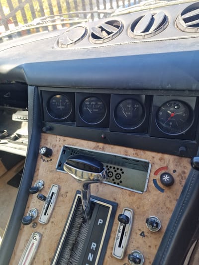 The image displays the interior of a classic car, focusing on the dashboard and center console from a slightly elevated, tilted perspective. No people are visible.
On the left, a vertical stack of four circular air vents is visible, with a glimpse of the outdoor environment through the car's front window, suggesting a sunny day. Adjacent to the vents is a black panel housing four analog gauges:
1. A clock, labeled "VEG QUARZO", indicating approximately 12:45.
2. A fuel gauge, labeled "FUEL", showing the tank is slightly over half full.
3. An oil temperature gauge, labeled "OIL TEMP.", with markings "140 210 280 F", and the needle resting around 140 F.
4. An ammeter, labeled "AMPERE", with markings "-40 0 +40", showing a reading near 0.
To the right, the center console features a wood-grain finish. Notable objects include:
* An air conditioning/heating control knob with a snowflake symbol and red/blue indicators.
* An empty slot, possibly for a car radio or stereo, exposing internal components.
* A prominent automatic transmission shifter with a chrome base and black knob, displaying "P R N D" for Park, Reverse, Neutral, Drive.
* Below the shifter, a slider control marked "0 F" and several smaller rotary knobs.
* A slim horizontal gauge or control labeled "Gs" towards the bottom right.
The interior shows signs of wear and age, including dust on the surfaces and scratches on the wood-grain. The overall setting is the cockpit of what appears to be a vintage vehicle, likely stationary during the day.