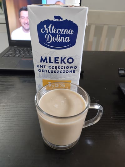 In Mieszkowice, Poland, a still life photograph captures a glass mug filled with a pale, creamy liquid, likely milk, placed on a dark, wooden surface. To the left of the mug, a portion of a white and blue carton of "MLEKO UHT CZĘŚCIOWO ODTŁUSZCZONE" (milk UHT partially skimmed) is visible, with Polish text and a drawing of a cow on a hill. The carton's branding, "Mleczna Dolina," is prominent. In the background, a laptop screen displays a man with a white shirt, smiling. The overall scene suggests a moment of personal time, possibly work or leisure, at a desk.
