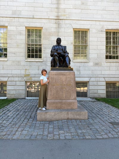 The image depicts a woman standing in front of the John Harvard statue located in Harvard Yard, Cambridge, Massachusetts, United States of America. The statue, cast in bronze, shows a seated figure wearing academic robes and holding a book. It rests on a granite pedestal inscribed with "JOHN HARVARD" and "FOUNDER". The woman, with short brown hair and wearing glasses, is dressed in a white t-shirt and a flowing olive green skirt, with white sneakers. She is smiling and looking towards the viewer, with her hands resting on the pedestal. Behind the statue and the woman is a large, imposing building constructed of light-colored stone blocks with evenly spaced, multi-paned windows. The ground in front of the statue is paved with cobblestones, and there is a strip of green grass to the right. The scene is illuminated by natural daylight, suggesting it is daytime.