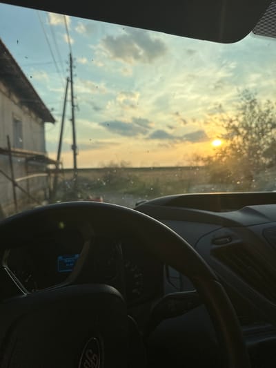 The image is taken from inside a vehicle, looking out through the windshield at a sunset. The view shows a rural landscape with a house on the left and a power pole. The dashboard and steering wheel of the vehicle are visible in the foreground. The sun is low on the horizon, casting a warm glow over the scene. The sky is filled with clouds. There are some dust or water droplets on the windshield, obscuring the view slightly. The car's interior features are dark, but a digital display on the dashboard shows the number "31". The steering wheel has a "Ford" logo on it. The location is identified as Mlekarevo, Bulgaria. There are no people visible in the image.