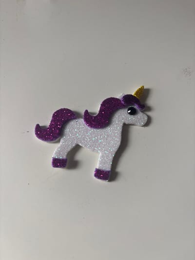The image shows a glittery white unicorn sticker with purple mane and tail, and a yellow horn. The unicorn sticker is lying on a white surface. The image was taken on a plain white surface, which could be a table or floor. There is no indication of the location in the city of Sauðárkrókur, Iceland.