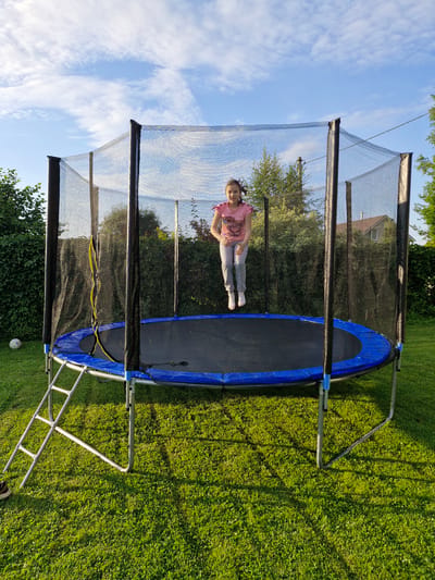 A young girl is captured mid-jump on a trampoline in a grassy backyard. She is wearing a pink t-shirt and grey pants. The trampoline has a safety net enclosure. The scene appears to be in a residential area with trees and houses visible in the background. The lighting suggests it is daytime. The provided crops do not offer any additional information regarding the specific location within Sorochkino, Russia.