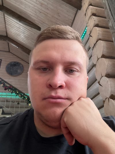 The image presents a close-up portrait of an adult male within an indoor setting in Matveevskoe, Russia. The subject, positioned centrally, has fair skin, short light-brown hair, and exhibits light facial stubble. He is wearing a dark, short-sleeved t-shirt and is observed resting his left hand beneath his chin. The background displays a rustic interior constructed predominantly of wood. To the subject's right, a wall composed of stacked horizontal logs is visible, featuring a vertical strip of green LED lighting. To the subject's left, a wooden ceiling with exposed beams and planking is present. A circular, dark-toned decorative object is mounted on the wall in the upper-left quadrant. Below this, a wooden railing with vertical balusters is adorned with a festive garland incorporating red and green spherical ornaments and green string lights. The overall lighting is consistent with an interior environment.