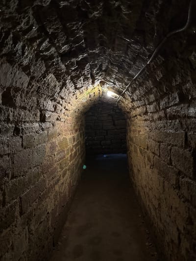 This media file shows a low-angle, wide shot of a dimly lit, narrow, brick-lined tunnel. The tunnel appears to be constructed of rough-hewn stones and bricks, with evidence of dampness and possibly water on the surfaces. A single, exposed light bulb illuminates the immediate foreground, casting stark shadows and highlighting the textured, irregular surfaces of the walls and floor. A metal conduit carrying electrical wiring runs along the left wall, disappearing into the darkness ahead.
The perspective draws the viewer into the tunnel, emphasizing its confined nature and the rough, aged construction. The lighting creates a sense of mystery and perhaps slight unease, as the tunnel recedes into deep shadow. The floor is paved with bricks, also appearing wet and worn. The overall impression is one of a historic or underground passage, possibly a cellar, service tunnel, or part of an older infrastructure. There is no indication of people or specific activity, suggesting a static, architectural capture. The setting is implied to be Posyolok Imeni Morozova, Russia, but this context is not visually evident within the image itself. There is no visible text.