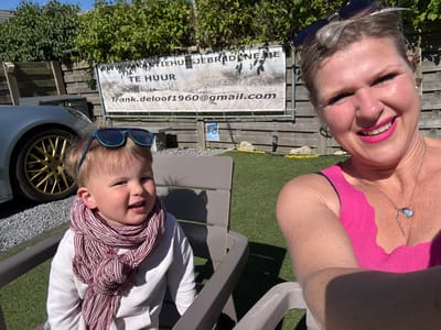 An outdoor, sunlit selfie captures an adult woman and a young child, likely in a backyard or patio area in Bredene, Belgium. The woman, on the right, smiles broadly at the camera, wearing a bright pink tank top and a silver necklace with a heart pendant. Her blonde hair is pulled back, and sunglasses rest on top of her head. On the left, a child, likely a toddler, sits in a grey plastic outdoor chair, also smiling. The child has blonde hair, sunglasses perched on top of the head, and wears a white long-sleeve top with a maroon and white striped scarf tied around the neck.
Behind the subjects, a wooden fence is visible, topped with green bushes and foliage. A portion of a silver car with a distinctive gold-colored wheel is seen on the left. The ground is covered in green artificial turf. Bright sunlight casts shadows, indicating it is mid-day or early afternoon on a clear, sunny day.
Attached to the wooden fence is a white banner. Visible text on the banner includes "WWW.VAKANTIEHUISJEBREDENE.BE", "TE HUUR" (Dutch for "FOR RENT"), and the email address "frank.deloof1960@gmail.com". The banner advertises a holiday home, consistent with the provided location context of Bredene, Belgium. Both individuals appear happy and relaxed, enjoying the sunny weather.