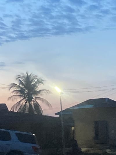 The image, taken in Amaseri, Nigeria, captures an outdoor scene at either dusk or dawn, characterized by a mix of natural elements and urban infrastructure. The sky dominates the upper and left portions of the frame, displaying a gradient from light blue with soft, scattered clouds on the far left to a paler, more diffused light towards the right, indicating low light conditions.
A brightly illuminated street light stands prominently in the mid-right, its lamp casting a strong glow against the dim sky. Below and slightly to the right of the street light, a large palm tree, likely a coconut palm, is silhouetted darkly, its dense fronds fanning out. Several thin power lines or communication cables are visible stretching across the sky, passing near the street light.
On the far right side of the image, parts of a dark, indistinct building or structure are visible, suggesting a house or commercial establishment. At the bottom right, partially obscured behind the palm tree, is a section of a white vehicle, possibly a van or SUV, with what appears to be a spare tire mount on its rear. No people are visible, and there is no discernible text in the frame. The scene presents a typical streetscape in a tropical setting.