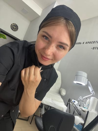 A woman wearing a black medical cap and black scrubs is taking a selfie in what appears to be a beauty salon or medical office. She is holding the collar of her scrub top with her right hand. Her left arm is bent and resting against her chest. In the background, there is a white dental chair covered in plastic, a desk with various beauty products and equipment, including a magnifying lamp, and a clock on the wall. The room is lit with artificial lighting. The text on the wall in Cyrillic is visible, which translates to "love for beauty is...". The image does not provide any specific location information within Stepnogorsk city administration, Kazakhstan.
