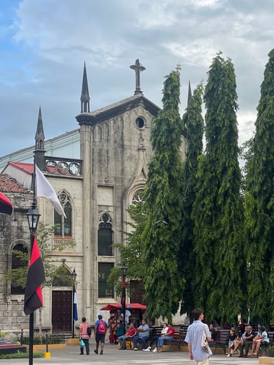 The image captures the facade of a stone church with pointed spires and a prominent cross on its roof, set against a cloudy sky. In the foreground, several tall, dense green trees obscure parts of the church. In front of the church, a paved plaza is visible with people walking and sitting on benches. A black and red flag is prominently displayed on a pole in the left foreground, next to a lamppost and a white flag. To the right, several market stalls with red awnings are set up. The scene appears to be in León, Nicaragua, likely near the Cathedral of León. People are casually dressed, and the atmosphere seems to be that of a public gathering space in a city.