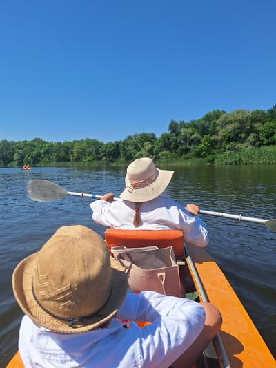 This image captures a serene moment on the river in the city of Potoky, Ukraine, where two individuals are enjoying a kayaking excursion. The perspective is from the rear of the kayak, looking forward. The sky is a clear, vibrant blue, devoid of clouds. On the water, the surface reflects the light, creating ripples and a sense of movement.
In the foreground, the back of two people is visible, both wearing white shirts and straw hats. The person in the front has a tan, woven hat with a black band. The person behind them wears a wider-brimmed straw hat with a decorative weave and a ribbon detail. This individual is actively engaged in paddling, holding a kayak paddle with a silver blade and a gray shaft. The paddle is positioned across the kayak, with its blade submerged in the water to the left. An orange seat and a light-colored tote bag are visible in the kayak.
In the mid-ground, the river extends towards a lush green shoreline. A line of trees forms a dense canopy, interspersed with reeds along the water's edge. Further in the distance, another kayak, occupied by two individuals, can be seen on the water, adding a sense of shared activity on the river. The overall scene is one of peaceful outdoor recreation, bathed in the bright light of a sunny day.