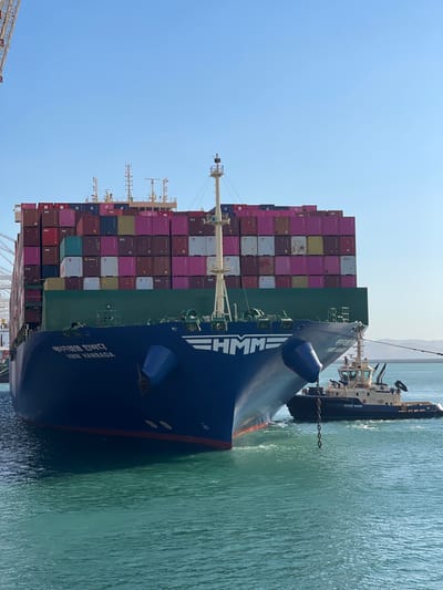 In Ksar al Majaz, Morocco, a large container ship named "HMM HANBADA" is being assisted by a tugboat in a body of turquoise water. The container ship is laden with numerous multi-colored shipping containers, stacked high. The tugboat, identified as "SVITZER TANGER," is positioned to the right of the container ship's bow and is attached by a chain, indicating it is actively involved in maneuvering the larger vessel. The sky is clear and blue, with distant hills visible on the horizon. The scene suggests port operations are underway.