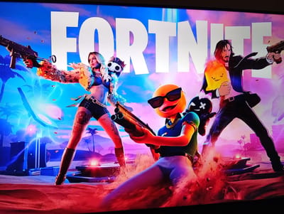 The media file is a digital image displaying the title screen for the video game Fortnite. The background features a vibrant, stylized tropical landscape with palm trees and a gradient sky transitioning from blue to purple, suggesting either dawn or dusk.

The foreground is dominated by three characters in action poses. On the left, a woman with blonde hair, wearing a fur-trimmed top and shorts, holds a submachine gun. Bullet casings are scattered around her, indicating an active combat scenario. In the center, a yellow, anthropomorphic duck-like character with sunglasses holds a shotgun. This character appears to be breaking through the ground, kicking up red dust and debris. To the right, a character resembling John Wick from the film series, dressed in a black suit, holds a pistol and a flaming object.

The overall tone of the image is energetic and action-packed, typical of a battle royale game. The scene suggests intense combat within a tropical environment. The prominent white text "FORTNITE" is displayed across the top of the image.