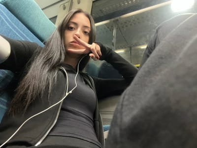 A female individual is depicted inside a train carriage, taking a selfie. She has long, dark hair and is wearing a black long-sleeved top with an unzipped black hooded jacket. White wired earphones are visible around her neck and chest. Her right hand is raised, forming a V-sign with her index and middle fingers; her fingernails are painted red. She looks directly at the camera with a neutral expression. To her left, part of a blue patterned seat is visible. To her right, a window reflects interior lighting and shows indistinct external lights, suggesting a station platform or sheltered area. The interior of the carriage features overhead lighting and visible structural elements. The specific location within the city of Adur, United Kingdom, cannot be determined from the visual information in the image.