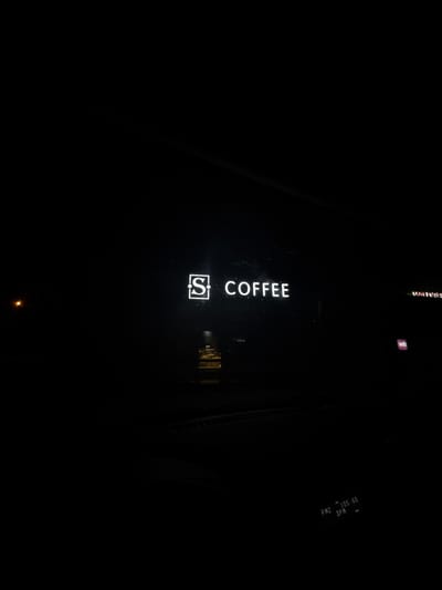 The image shows a nighttime scene with a bright white neon sign that reads "S COFFEE". The sign is located on a dark building facade. To the right of the "S COFFEE" sign, there is a smaller, purple neon sign with the letters "WB" inside a square. Further to the right, another neon sign is partially visible with faint white lettering. In the lower right corner, a digital display shows "FM2 105.60 DFM". The image is taken from a low angle, possibly from a car, as indicated by the presence of a dashboard reflection at the bottom. The overall environment is dark, suggesting it is late at night. There is no information in the image to suggest the location is in Lubnya, Russia.
