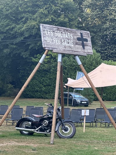 In the city of Saint-Vaast-du-Val, France, an outdoor gathering is taking place. A wooden sign with the inscription "LES SOLDATS DU FEU SACRE" and a black cross is prominently displayed, supported by three wooden poles. In front of the sign, a black motorcycle is parked on a grassy area. Behind the motorcycle, rows of black chairs are arranged, suggesting an audience or seating area. A dark-colored car is visible in the background, partially obscured by a large tent with a light-colored canopy. The environment is characterized by lush green trees and foliage.