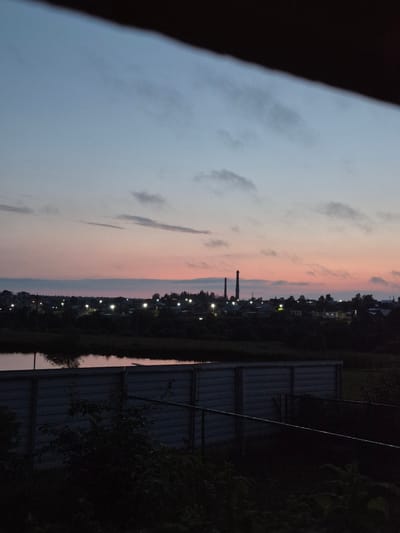 The image captures a twilight scene in Sars, Russia. The sky displays a soft gradient of colors, transitioning from a pale blue at the horizon to delicate pink and gray hues higher up, with scattered clouds. The foreground is dominated by a dark, out-of-focus area on the left, possibly a railing or other foreground object.
To the right, a long, corrugated metal fence runs vertically along the frame. Beyond the fence, silhouetted against the colorful sky, is a collection of buildings and trees, suggesting a residential or semi-rural area. Several streetlights are visible, casting pools of yellow light along the treeline, indicating that dusk is settling. Two tall, industrial chimneys stand out prominently in the middle ground. The overall atmosphere is serene and quiet, typical of the evening hours. The lighting suggests it is either dawn or dusk, given the pastel colors in the sky. There is no visible text in the image.