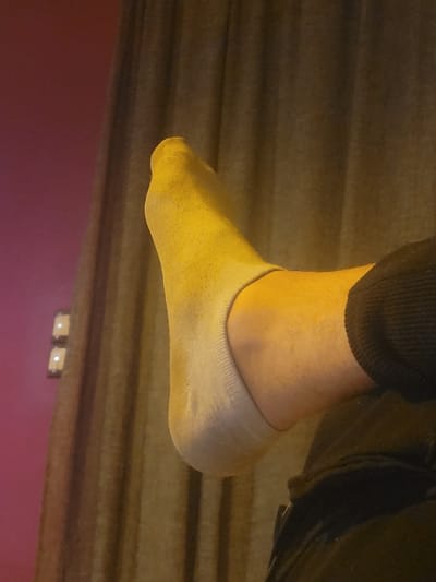 The image displays a human left foot, elevated and seen in profile, within a domestic setting in Ανδραβίδα, Greece. The foot is covered by a light-colored, low-cut ankle sock, with the lower part of a dark garment, likely trousers, visible on the leg. The background consists of a solid magenta-colored wall on the left, meeting a dark brown or charcoal curtain with vertical folds on the right. Two light-colored, square fixtures, appearing to be electrical switches or outlets with illuminated indicators, are mounted on the magenta wall near the curtain. A warm, yellowish light illuminates the foot, contrasting with the darker background.