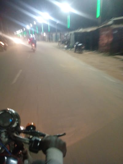 This image shows a nighttime scene on a street in Tundun Wada West, Nigeria. The perspective is from someone riding a motorcycle, with the handlebars and part of the dashboard visible in the foreground. The road appears to be paved with a sandy or dirt shoulder. Along the right side of the road, there are low-rise buildings with dark, possibly tin roofs. Several streetlights are illuminated, casting bright white light, and there are also some vertical green lights visible, possibly decorations or additional lighting.

In the distance, there are other vehicles, including at least one motorcycle with a rider. The lighting suggests it is evening or night, and there are no immediate cues about the weather other than the darkness. The overall impression is of a busy, illuminated street at night, perhaps during a local event or simply a regularly lit thoroughfare. The motion blur indicates movement, consistent with being on a moving motorcycle. No text is visible in the image. The scene is somewhat blurry due to the low light conditions and the movement.