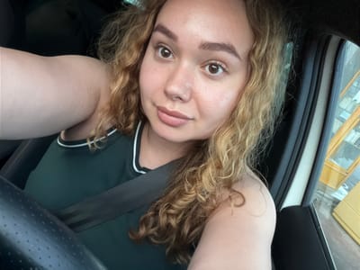 A close-up selfie, originally rotated 90 degrees clockwise, features a young woman inside a vehicle during the daytime. She has fair skin, striking brown eyes, full lips, and well-defined eyebrows. Her long, voluminous, curly hair, a blend of blonde and light brown, cascades around her face. A notable detail is a heart-shaped curl of hair near her left temple. She wears a dark green or teal sleeveless top with white stripes accentuating the trim.
The setting is clearly a car interior, evidenced by a black seatbelt diagonally crossing her chest. Parts of the vehicle's interior are visible, including a dark steering wheel on the right side and a perforated ceiling panel in the upper right corner. Through the window on the left, a building with yellow or tan siding and dark windows can be seen, suggesting an urban or suburban outdoor environment. The lighting implies it is daytime. The woman has a neutral expression, looking directly into the camera. There is no visible text.