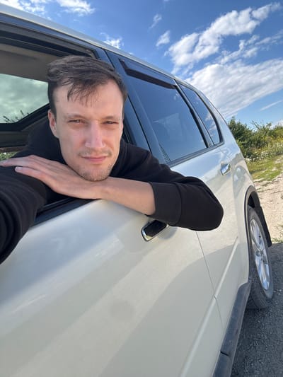 A young adult male with light brown hair and a stubble beard leans his head on his arm, which is resting on the open window of a white SUV. He is wearing a black hooded sweatshirt. The car is parked on a gravel road with sparse vegetation and trees in the background. The sky is blue with scattered white clouds. The image appears to be a selfie taken outdoors in a rural or semi-rural area, potentially near Abrykosivka, Ukraine.