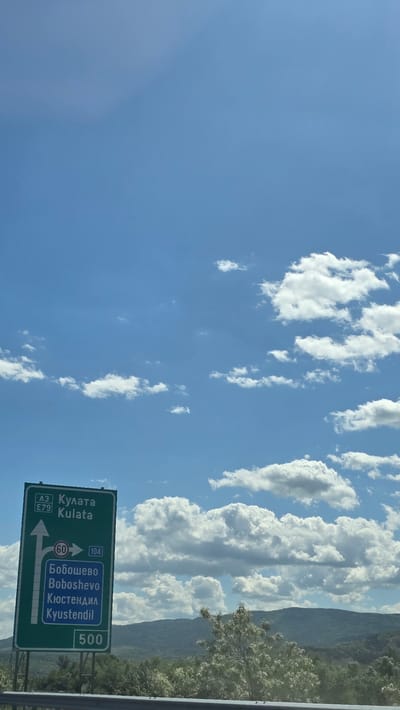 The image depicts a daytime scene on a highway in Bulgaria, near the city of Blajievo. Dominating the view is a blue sky with scattered white cumulus clouds. A green highway sign is visible, indicating directions to: "Кулата Kulata" (left), "Бобошево Boboshevo" (straight), and "Кюстендил Kyustendil" (straight). The highway numbers are A3 and E79 for Kulata, and 104 for Boboshevo and Kyustendil. A speed limit sign of 60 is visible above the directional arrow. The sign also indicates that Kyustendil is 500 kilometers away. In the background, there is a mountain range with vegetation. The image appears to be taken from inside a vehicle.