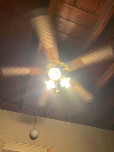 A low-angle, wide shot captures a ceiling fan with its blades blurred from rotation, indicating it is in operation. The fan is mounted to a wooden-paneled ceiling, with exposed wooden beams also visible. The fan has a brass-colored housing and features at least three lights, which are illuminated and contribute to the bright glare in the image. The overall setting appears to be an interior room with a rustic, wooden aesthetic. To the right of the fan, a portion of a light-colored wall is visible, with a simple, globe-shaped light fixture hanging from a thin cord. The scene is illuminated by the fan's lights and possibly ambient light from the nearby wall fixture. The image provides no clues about the time of day or weather conditions. No people are visible in the frame. There is no discernible text within the image.