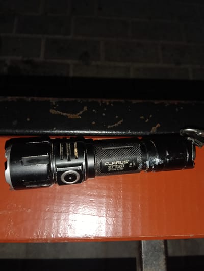 The image depicts a black flashlight lying on an orange surface. The flashlight has a textured grip and a lens at one end. There are some markings on the flashlight body, including the brand "KLARUS" and the model "XT11GT". A key is attached to the flashlight via a ring. The background appears to be a brick wall. The setting does not provide any specific information to determine its location in Florești, Moldova.
