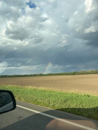 The image depicts a rural landscape with a plowed field stretching towards a dense line of trees under a dramatic, cloudy sky. A faint rainbow is visible in the distance, arcing across the sky. In the foreground, a car's side mirror is visible on the left, reflecting the sky and a portion of the road. The road itself, with a white line marking its edge, occupies the lower part of the frame. Green grass borders the field and the road. The scene appears to be located in the vicinity of Polivyanka, Russia, suggesting a transitional weather event with a potential for rain. There are no people or specific actions captured in the image. The overall impression is one of natural beauty and the transient effects of weather.