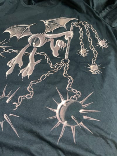 The image depicts a dark-colored fabric with a graphic design printed on it. The design features a cartoonish, bat-winged creature wielding a flail. The flail is comprised of a chain connected to several spiked balls of varying sizes. The creature appears to be in motion, possibly swinging the flail. The fabric seems to be part of a piece of clothing, potentially a t-shirt. There is no information in the image to suggest a location in La Lucila, Argentina.