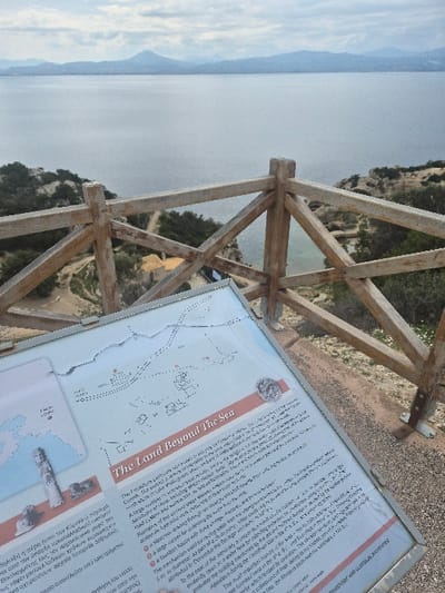 An elevated, downward-angled view in the Municipal Unit of Loutraki - Perachora, Greece, captures a coastal landscape under an overcast sky. In the foreground, an informational sign with a light-colored background is angled towards the viewer, featuring text in Greek and English, including a section titled "The Land Beyond The Sea," a map, and pictorial elements. A light-colored wooden railing with diagonal cross-braces extends across the mid-ground, separating the immediate vantage point from the lower coastal terrain. Below the railing, a rugged, sparsely vegetated coastline with rocky outcrops descends towards a body of water. A narrow dirt path is visible, winding through the terrain, leading towards a small bay where a light-colored building with a yellow roof and a partially visible Greek flag are situated near the water's edge. The background is dominated by a vast, calm expanse of grey-blue water, with a distant range of mountains or hills visible on the far horizon beneath a cloudy sky. No individuals are present in the scene.