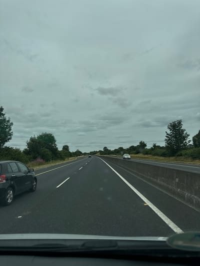 An overcast sky looms over a multi-lane highway in Ballyduff South, Ireland. The road is paved with dark asphalt, marked by white lines. A black hatchback is in the left lane, closest to the viewer. In the right lane, a white SUV is visible further down the road. The sides of the highway are lined with green vegetation and trees. A concrete barrier separates the lanes of traffic. The scene suggests a typical day of travel on a major road in the Irish countryside.