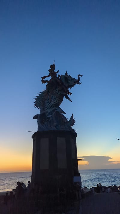 The image captures a statue of a Barong at sunset in Pererenan, Indonesia. The statue depicts a mythical creature, and is mounted on a pillar. The sky is blue, transitioning to orange as the sun sets over the ocean. The sea is visible in the distance, with people seen on the beach.