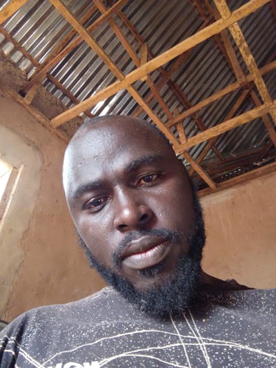 The image is a low-angle self-portrait of a dark-skinned, bald man with a full, dark beard. The background reveals a simple interior space. The ceiling is constructed of corrugated metal sheets supported by a wooden frame. The walls are light brown. A window is visible on the left. The shirt the man is wearing is dark with a light-colored pattern. The specific location is Mulke Barikin Sayejo, Nigeria.