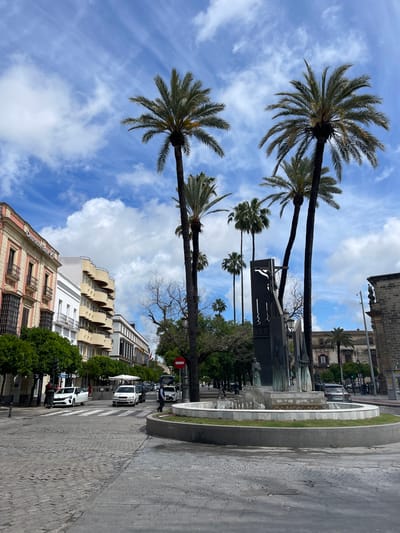 This street scene in Jerez De La Frontera, Spain, features palm trees lining a thoroughfare on a sunny day with a blue sky marked with puffy white clouds. The street is paved with cobblestones and includes a zebra crosswalk where a man is crossing. Adjacent to the crosswalk, cars and other vehicles are parked along the side of the road. Buildings line the side of the street, including a multistory building with arched balconies. In the center of the street, a modern sculpture stands next to a tiered fountain with a green border. A 'Do Not Enter' sign is visible at the intersection of the fountain and the street. Several other people can be observed in the background near the fountain and buildings.