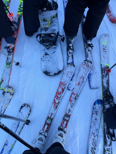 The image, captured from an overhead perspective, showcases a collection of winter sports equipment and parts of individuals on a snow-covered surface, likely on a mountain in Courmayeur, Italy. The scene primarily features skis, snowboards, ski boots, and ski poles, all partially covered in fresh snow.

Multiple people are implied, with their legs clad in black snow pants, and their feet secured in various bindings. In the upper left, a pair of black boots are attached to skis. Another pair of skis prominently displays the brand "FISCHER" and model details such as "RC4" or "RCR," featuring red, white, and black designs, with white ski boots visible. These skis are positioned near another pair of black ski poles and a partial view of a ski boot.

Toward the center, a black snowboard with black bindings, also dusted with snow, is visible. It features yellow and black graphics, with some indistinct text that could be "STK" or "SOL". Nearby, the lower legs and boots of a person using this snowboard are visible. Another set of skis with yellow, black, and white patterns can be seen in the lower left, accompanied by ski poles. At the top of the frame, another pair of skis shows partial text "4F" and potentially "HEAD."

The entire setting is dominated by bright white snow, indicating cold weather and potentially recent snowfall. The even lighting suggests it is daytime, possibly under an overcast sky, as there are no distinct shadows. The arrangement of the gear suggests a group has gathered, perhaps taking a break or waiting, during their skiing and snowboarding activities on a resort slope.