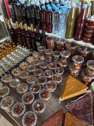 This is a high-angle, full-shot, eye-level, indoor photograph of a market stall displaying various food and drink items. The stall is located in Aranisi, Georgia. In the background, numerous bottles of wine and other beverages are arranged on shelves, with some bottles on display in the foreground as well. To the right of the bottles, there are stacks of small, clear plastic containers filled with nuts and dried fruits. In the foreground, several large, flat, triangular pieces of fruit leather are wrapped in plastic. The overall impression is that of a well-stocked and diverse market stall offering a variety of traditional Georgian products. There are no people visible in the image. The lighting is natural, suggesting the stall is open to sunlight. The surfaces of the stall appear to be wooden.