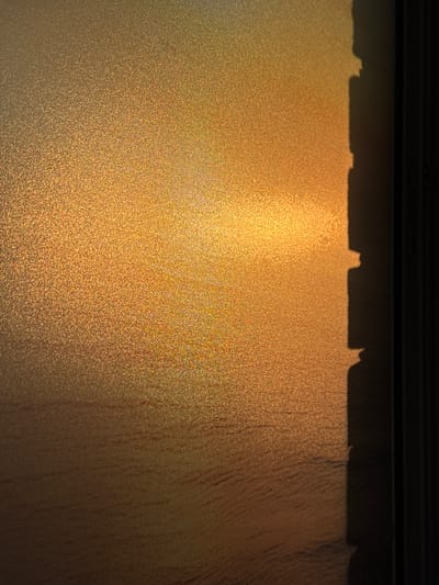 The image shows a frosted window, partially illuminated by golden sunlight. The top of the window is shadowed by a dark frame and what appears to be the edge of a building or overhanging structure, creating an irregular shadow pattern. The frosting on the glass obscures the view outside, giving a textured appearance to the light filtering through. The right side of the window is darker, suggesting a difference in light exposure or the presence of an object casting a shadow.
It is not possible to verify the location of this image in Kladovitsy, Russia, from the provided details.