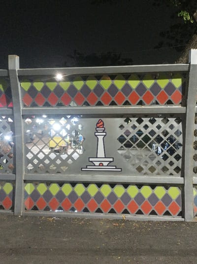 A nighttime photograph depicts a section of a decorative metal barrier or fence, likely situated in the Special Capital Region of Jakarta, Indonesia. The fence is composed of gray vertical posts and horizontal beams, featuring three distinct horizontal panels. The top and bottom panels display a repetitive pattern of interlocking diamond shapes in lime green, muted purple, and terracotta orange. The central panel is a lighter gray metal with a grid of small, open diamond-shaped perforations. Centered on this middle panel is a white silhouette of the Monas (National Monument) with a red flame at its apex, outlined in black, serving as an emblem of Jakarta. Bright, white artificial lights from an indistinct source are visible through the perforations, reflecting off the metal and indicating an illuminated area or activity beyond the fence. The foreground consists of a dark asphalt surface with minimal scattered debris. No individuals or specific actions are discernible within the frame.