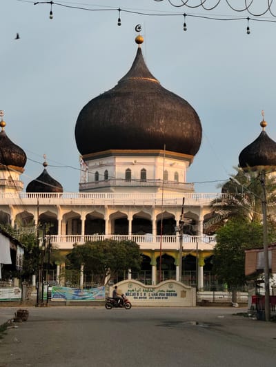 The image depicts the exterior of the Masjid B.S.R. L. Khairifah Ibrahim mosque, located in Pulodolang, Indonesia. The mosque features multiple domes, with a large, dark, conical central dome prominently displayed. Smaller domes are visible on either side, and a smaller, pointed dome is situated on the main structure. The building has a white facade with arched colonnades on the lower level and a white balustrade surrounding the upper level. Several windows are visible on the upper floors.
In the foreground, a paved road is present, with a person riding a red motorcycle moving from left to right across the frame. To the left of the road, there is a woven basket on the ground and some debris. Further back, a white sign with Arabic and Latin script identifies the mosque and its location: "MASJID B.S.R. L. KHAIRIFAH IBRAHIM KEC. MATANG KUI, ACEH UTAR". Banners are displayed behind the sign. Lush green trees and palm trees surround the mosque. Overhead, a string of bare light bulbs is suspended by wires. The sky is a pale blue, with a single bird visible in flight on the left side of the image. The lighting suggests it is either early morning or late afternoon.