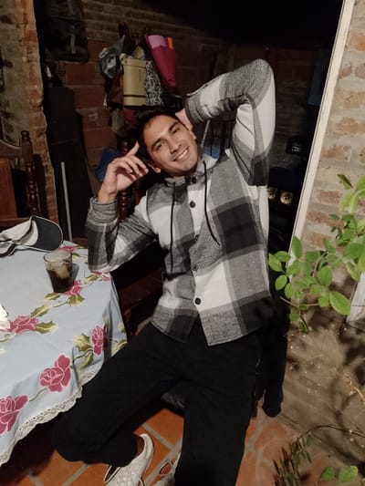 A young adult male is sitting at a table outdoors, smiling and posing for the camera. He is wearing a black and white plaid hooded shirt, black pants, and white sneakers. His arms are raised behind his head, and one hand is near his hair. On the table is a floral patterned tablecloth, a glass with a dark beverage and ice, and a baseball cap. The background is a brick wall and a dark, cluttered area, possibly a storage space. A plant is visible near the right side of the frame. The location is likely Gobernador Julio A. Costa, Argentina.