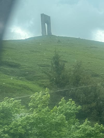 The image, appearing to be captured with a vertical orientation, displays a expansive green, grassy hillside stretching towards an overcast sky. Perched atop the hill is a prominent, arch-like concrete or stone structure, recognized as the Arch of Liberty (Svoboda Arch) on Gorni Vrah peak, near Hristo Danovo, Bulgaria. This monument features a large, open archway and commemorates the Battle of Hristo Danovo during the Russo-Turkish War.
In the foreground, on the right side, dense green trees and foliage are visible, partially framing the view of the hill and monument. Other scattered trees and bushes dot the verdant slope. The sky is predominantly covered by light-grey, dense clouds, indicative of an overcast or hazy day, with some brighter areas suggesting the sun is obscured.
No people are present, and no specific activity is taking place. The scene captures a peaceful, natural landscape featuring a significant historical landmark under muted daylight conditions. No discernible text is visible within the image.