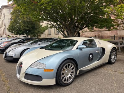 A line of high-performance Bugatti Veyron automobiles is parked on a paved outdoor surface in Molsheim, France. The leading vehicle, a Bugatti Veyron 16.4, is finished in a two-tone scheme of metallic light blue and cream-white. It displays a black circular event decal with the white numeral '51' on the driver's side door panel. Chrome multi-spoke wheels are fitted, with yellow brake calipers visible on the front wheel. Directly behind, a silver Bugatti Veyron is visible, bearing French registration plate 'W-391-DB', followed by additional luxury cars in black and orange.

The scene is set in an open area, likely a public square, in Molsheim. Background elements include several large deciduous trees with green foliage. To the left, a multi-story building with light-colored facades and multiple windows is present. To the right, a substantial historical building constructed of reddish-brown stone or brick, consistent with the architecture of the Église des Jésuites in Molsheim, is partially observed. A wooden picnic table and bench are positioned to the right rear of the foreground vehicle, adjacent to a dark metal fence. Distant figures are faintly visible among the vehicles on the left, one wearing a high-visibility vest.