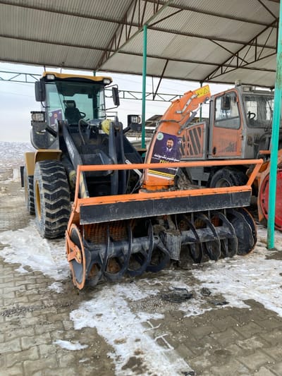 The image depicts a scene of heavy machinery parked under an open-sided shelter with a corrugated metal roof supported by green metal poles, located in Kan Mehtarzai, Pakistan. In the foreground, a large yellow wheel loader, bearing the identifier "LG936FN" on its lifting arm, is equipped with a prominent orange front-mounted snowblower attachment featuring a multi-bladed auger. Two identical political posters are visible; one affixed to the cab of the wheel loader and another to the orange snowblower. Each poster displays the image of a bearded man, alongside text in an unknown script and the acronyms "PDM" and "PDMA." Partially visible behind the snowblower is an older, orange-red utility vehicle with a cab. The ground directly beneath and around the machinery is paved with grey interlocking bricks, interspersed with melting snow, dirt, and gravel patches. The distant background reveals a barren, hilly landscape under an overcast sky. No individuals are present in the scene.