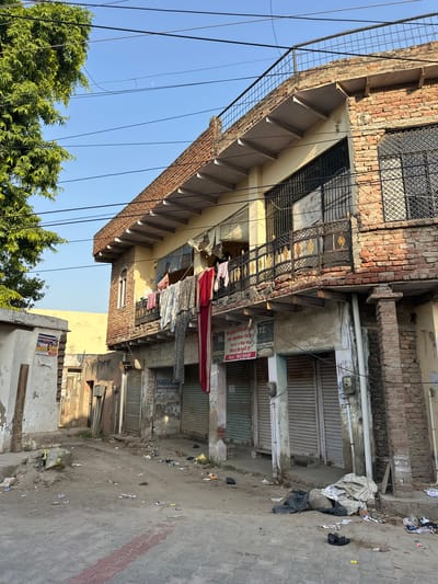 The image captures a street scene in Mathura, India. The buildings lining the street are constructed primarily of brick and concrete, with some displaying metal security grills. Laundry lines strung between the buildings hold various garments, and a large red piece of fabric hangs prominently. The street is littered with trash and debris. A sign with Hindi text, reading "लेडीज ब्लाउज ले. सूट लहगा फ्री. यह ड्राईक्लीनिंग की की अब महाविद्या चौराहा 59-M, 11-फेस पर शिष्ट हो चुकी है। Mob.: $897564937" can be seen on one of the buildings. The sky is a clear blue. A tree with dense green foliage is visible in the lower left corner.
