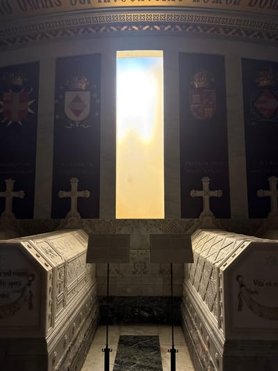The image depicts the Royal Pantheon of the Monastery of San Lorenzo de El Escorial, located in San Lorenzo de El Escorial, Spain. The scene is dimly lit, with a bright rectangular window at the back of the chamber casting a strong backlight. In the foreground, two ornate sarcophagi are visible, made of white marble and decorated with intricate carvings. The sarcophagus on the left has text inscribed on its side, reading "er sed vivam, obo opera minini." The sarcophagus on the right has text that reads "Vita ex m".

Behind the sarcophagi, on the walls, are four dark panels, each featuring a royal coat of arms and a crown at the top. The names "PHILIBERTVS", "MARGARITA", "FERDINANDVS", and "MARIA" are inscribed on these panels, along with their familial relationships. Between these panels are two large white crosses, also carved from marble, mounted on ornate bases. The floor appears to be made of polished dark stone with lighter marble patterns. Two metal stands with informational plaques are positioned between the sarcophagi. The overall atmosphere is solemn and monumental, characteristic of a royal burial site.